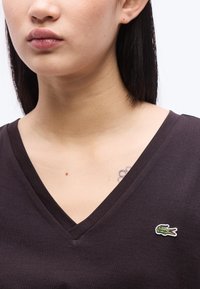 Femme aux longs cheveux noirs portant un t-shirt noir à col en V avec un logo de crocodile vert sur la poitrine et un petit tatouage de fleur près de la clavicule.