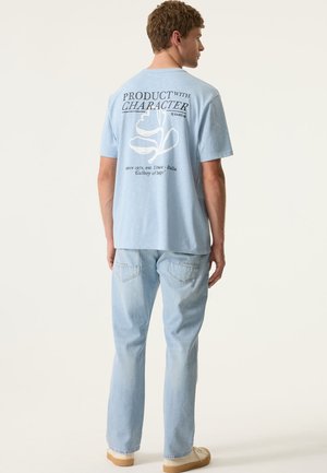 Lichtblauw katoenen T-shirt met witte grafische tekst en bladontwerp op de achterkant, gecombineerd met lichtblauwe spijkerbroek en bruine schoenen.