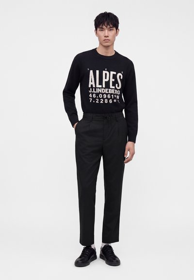 Jeune homme portant un pull noir avec un texte blanc "ALPES" et des coordonnées, pantalon noir, chaussettes blanches et baskets noires, debout devant un fond blanc.