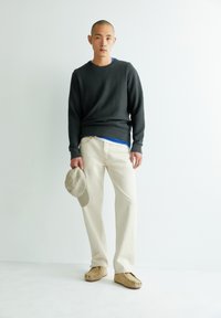 Pull en laine gris texturé, pantalon beige clair et chaussures slip-on beiges. Tient une casquette gris clair. L'arrière-plan est blanc uni.