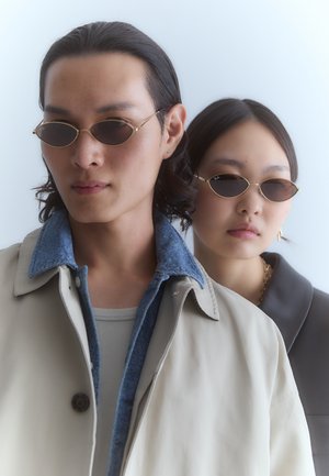 Aurinkolasit kullanenkehyksillä, joissa on soikeat linssit, päällä kahdella henkilöllä. Toinen on pukeutunut beigeen takkiin, jossa on farkuista tehty kaulus; toinen tummanväriseen kaulukselliseen puseroon.