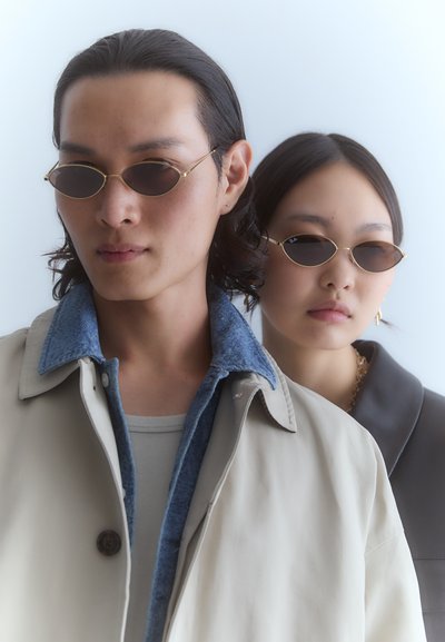 Des lunettes de soleil avec une monture en or et des verres ovales, portées par deux personnes. L'une porte un manteau beige avec un col en denim ; l'autre est vêtue d'un haut sombre avec col.