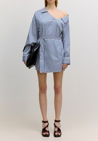 Gestreiftes Hemdkleid in Blau und Weiß mit schmalem Taillengürtel, langen Ärmeln und einem schulterfreien Design. Getragen mit einer schwarzen Handtasche und braunen Sandalen.