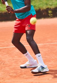 Tennis shoes featuring a white upper, green and yellow accents, coupled with red shorts and a blue shirt against a clay court backdrop.