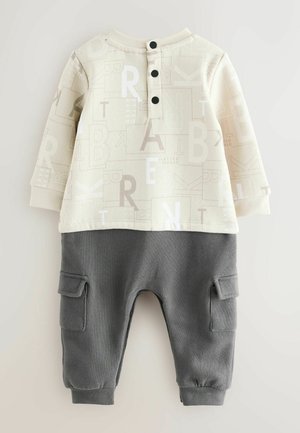 Két részből álló öltözet krémszínű felsőrésszel, amely betűmintákkal díszített, és szürke alsó résszel, amely béka zsebekkel rendelkezik. Az anyag puha, bordázott mandzsettákkal.