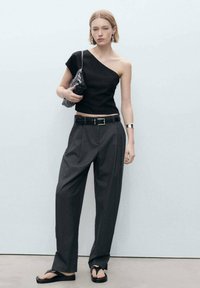 Top nero a spalla unica, pantaloni grigi a gamba larga, cintura nera con fibbia quadrata, sandali neri. Gli accessori includono una borsa nera strutturata.
