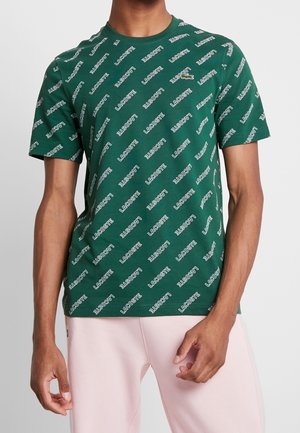 Persoon draagt een groen T-shirt met korte mouwen met herhaalde witte "Lacoste" tekst en een klein krokodillenlogo, gecombineerd met lichtroze broek.