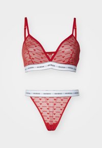 Set di lingerie rosso trasparente composto da un reggiseno triangolare con spalline sottili e un perizoma abbinato, entrambi decorati con motivi a forma di cuore e una banda bianca "GUESS".