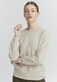 Beige hettegenser laget av mykt stoff, med rund halsutringning, ribbestrikkede ermer og en subtil X-søm detalj foran.