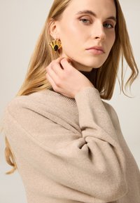 Kolczyki w kolorze złota o wachlarzowatym kształcie z czarnym kamieniem w centrum, noszone z beżowym, fakturowanym swetrem. Widoczny gładki detal tkaniny.