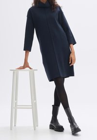 Robe à manches longues bleu marine avec un col bénitier, en tissu doux. Associée à des bottines noires et des collants noirs, debout à côté d'un tabouret blanc.