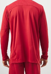Rød langærmet atletisk skjorte med teksturerede skulderpaneler, en glat krop og en afslappet pasform. Letvægtsstof velegnet til sport.