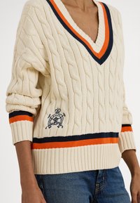 Kremasti džemper pletenog uzorka s V-izrezom, ukrašen mornarsko plavim i narančastim prugama na ovratniku i manžetama, s izvezenim logotipom na prednjem dijelu.