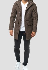 Brun uld-blandingsfrakke med hætte, toggleslås, forlommer og tekstureret overflade, båret over en hvid V-hals skjorte og sorte jeans.