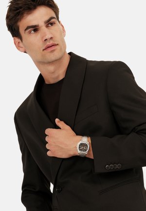 Sort blazer med sjalskrave, parret med en sort skjorte. Sølvur med sort urskive. Formelt tøj med knapdetalje på ærmerne.