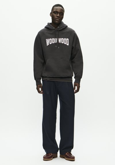 Siva kapucijska majica s logotipom "WOOD WOOD", kengurujskom žepom, u kombinaciji s modrim širokim hlačama i smeškim cipelama. Prazna pozadina.