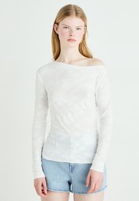 Hvit langermet topp med blomsterpreget mønster, off-shoulder design, laget av gjennomsiktig stoff, kombinert med denimshorts.