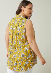 Blusa senza maniche color senape con un motivo floreale in bianco e nero, colletto a spalla e una vestibilità morbida con un orlo leggermente curvato.