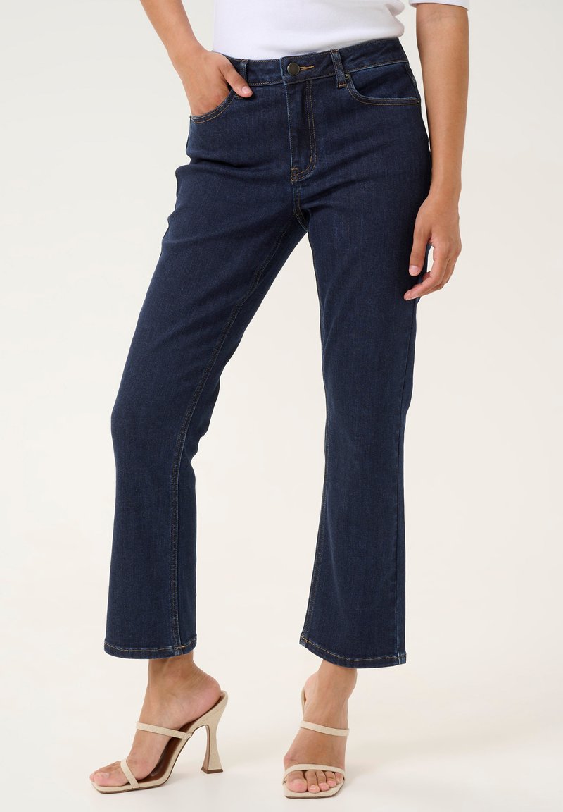 Mörkblå denimjeans med rak skärning, hög midja och synlig sömnad. Bärs med beigefärgade klacksandaler, vilket ger en avslappnad look.