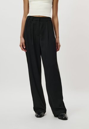 Vrouw draagt zwarte broek met hoge taille en brede pijpen, zwarte schoenen en een cropped witte mouwloze top tegen een effen achtergrond.