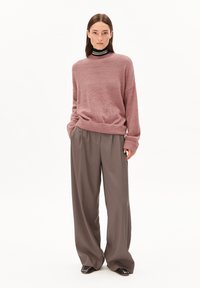 Eine Frau steht in einem übergroßen rosa Pullover, weiten grauen Hosen, einem schwarzen Rollkragenpullover mit weißen Streifen und schwarzen Schuhen mit Metallnägeln.