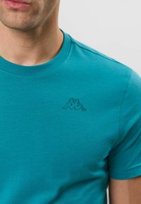 Homme portant un t-shirt bleu sarcelle à col rond avec un petit logo brodé de deux figures assises sur la partie supérieure gauche de la poitrine.
