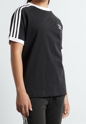 Osoba nosząca czarną koszulkę Adidas z białym kołnierzem, mankietami i trzema białymi paskami na ramionach.