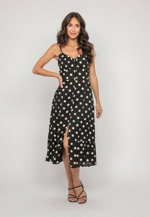 Femme debout et souriante, portant une robe noire sans manches à pois blancs et des sandales noires à brides sur un fond uni.