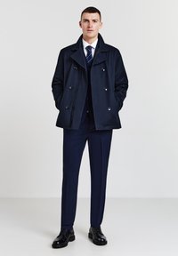 Cappotto peacoat blu navy con design doppiopetto e bottoni argentati, indossato sopra un completo e cravatta abbinati, completato da scarpe nere.