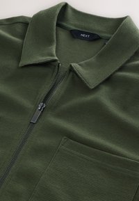 Chaqueta verde con cremallera, confeccionada en una tela texturizada, que incluye un cuello plano, cierre de cremallera y un bolsillo en el pecho como detalle adicional.
