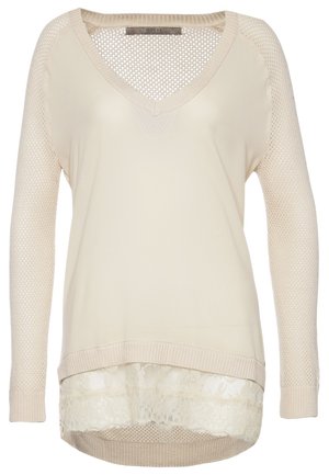 Femme portant un pull crème à col en V avec un ourlet en dentelle, un pantalon skinny noir, des bottines taupe et tenant une pochette beige.