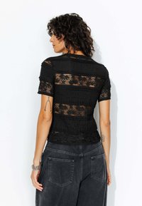 Haut en dentelle noire avec un tissu texturé à rayures horizontales, une silhouette cintrée et des manches courtes. Porté avec un bas en denim foncé, ample.