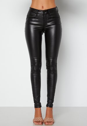 Femme portant un pantalon skinny en cuir noir taille haute et des talons ouverts tissés beige sur un fond clair uni.