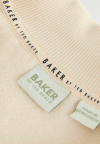 Tissu couleur crème avec une texture côtelée, arborant des étiquettes noires et vertes indiquant "BAKER by Ted Baker" et "Fabriqué au Bangladesh."