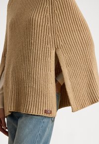 Paksu kudumine poncho soojas beežis värvitoonis, millel on külgmised lõhed ja väike nahklogoga silt allääres. Tekstuuriga kangas ribakujundusega.