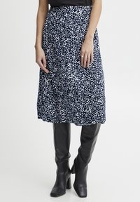 Žena v tmavě modré midi sukni s bílým leopardím potiskem a černými vysokými kozačkami až ke kolenům, stojící na jednobarevném pozadí.