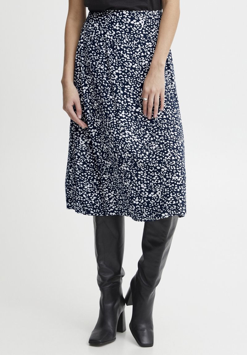 Nainen, jolla on päällä tummansininen midihame, jossa on valkoista leopardiprinttiä, ja mustat korkeat polvisukat, seisoo yksinkertaista taustaa vasten.