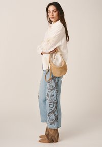 Femme en blouse blanche et jeans bleus brodés tenant un sac à main croissant couleur beige, portant des bottines beige en daim à franges, debout de profil.