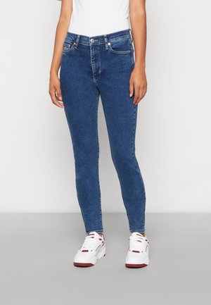 Femme portant un jean skinny bleu taille mi-haute et des baskets blanches avec des accents rouges, debout devant un fond clair uni.
