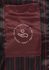 Bordeaux zorglabel op een zwarte stoffen achtergrond met dunne rode strepen, met daarin een icoon van een hand en een hart, en tekst over verzorgingsinstructies.