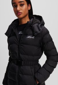 Chaleco puffer negro con cierre de cremallera, capucha y cintura con cinturón. Cuenta con secciones acolchadas y un logo en el pecho. Textura suave y lisa.