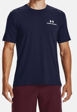Mężczyzna ubrany w granatową koszulkę Under Armour z krótkim rękawem i bordowe szorty, stojący z luźno opuszczonymi rękami wzdłuż ciała.