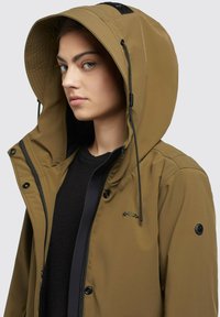 Giacca impermeabile khaki con un grande cappuccio regolabile, dettagli neri e zip frontale. Presenta bottoni a scatto e dettagli del logo sul petto.