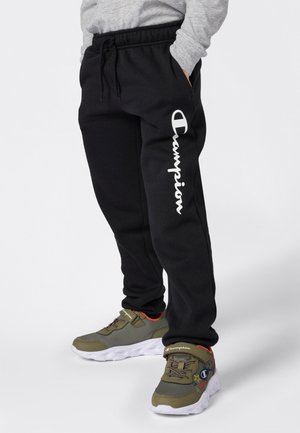 Persona vistiendo pantalones de chándal negros de Champion con texto del logo en blanco y zapatillas deportivas verde olivo con suelas blancas y detalles multicolores.