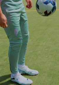 Lichtgroene sportbroek met een paarse logo en witte voetbalschoenen met zwarte accenten en roze noppen, staande op een groen veld.
