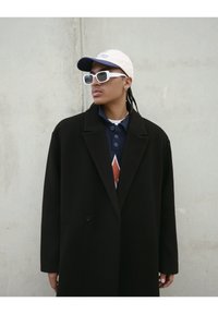 Manteau noir à coupe ajustée, porté sur un polo bleu marine avec un motif de losanges rouges. Une casquette blanche et des lunettes de soleil rectangulaires complètent le look.