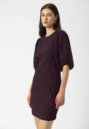 Junge Frau, die ein dunkelviolettes, strukturiertes Kleid mit gepufften, ellenbogenlangen Ärmeln trägt, steht vor einem schlichten weißen Hintergrund.