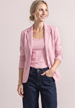 Donna sorridente con capelli biondi raccolti in uno chignon, che indossa un blazer rosa e una canottiera, jeans blu scuro, con una mano in tasca.