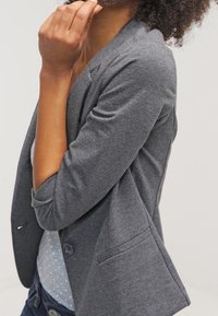 Blazer gris avec des revers crantés, une fermeture à bouton unique et deux poches avant. Porté sur un haut bleu clair à pois blancs.