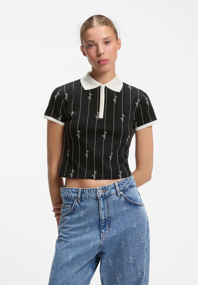 Jeune femme debout, les mains derrière le dos, portant un polo noir court avec un col blanc et un jean bleu taille haute.